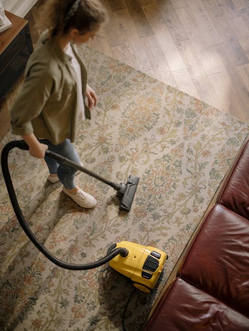 A woman is vacuuming a decorative, patterned area rug with shades of beige, green, and coral in a living room. She is holding a yellow vacuum cleaner and wearing casual clothing, including a beige jac
