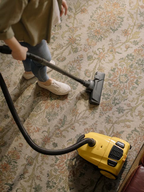 A woman is vacuuming a decorative, patterned area rug with shades of beige, green, and coral in a living room. She is holding a yellow vacuum cleaner and wearing casual clothing, including a beige jac